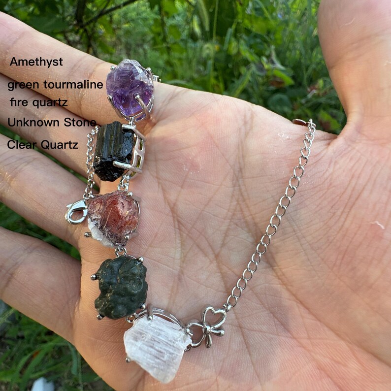 May include: A silver chain bracelet featuring various raw gemstones, including amethyst, green tourmaline, fire quartz, an unknown stone, and clear quartz. The bracelet is held in a hand against a blurred green background.