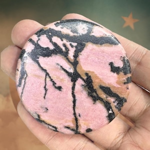 May include: A round, polished stone with a pink base color, featuring black and tan veining patterns. The stone is held in a hand, showcasing its natural textures and colors. The stone's surface is smooth and reflective.