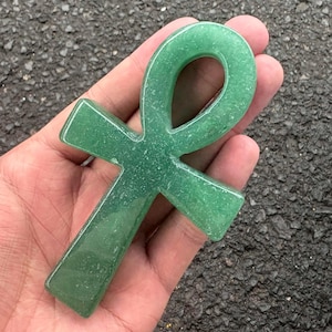 May include: A smooth, polished green aventurine stone carved into the shape of an ankh symbol. The object features a looped top section and a vertical base, displaying a consistent emerald green colour with subtle, natural sparkling mineral inclusions.