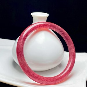 May include: A translucent, circular bangle bracelet crafted from a deep pink, crystalline material with a smooth, polished surface. The bangle features a consistent rounded profile and a vibrant, uniform reddish-pink hue throughout its structure.
