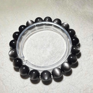 Könnte beinhalten: Ein kreisförmiges Armband aus glatten, kugelförmigen schwarzen Perlen mit einem reflektierenden, metallischen Glanz. Die Perlen sind in einer durchgehenden Schleife aufgereiht und zeigen eine einheitliche Farbpalette von Dunkelgrau bis Schwarz.