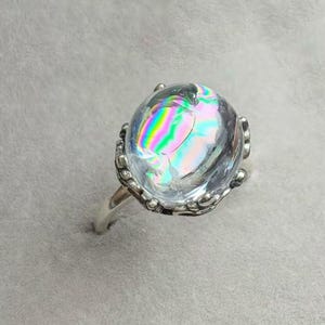 Könnte beinhalten: Ein silberner Ring mit einem großen, klaren, oval geformten Stein mit schillernden Regenbogenfarben. Der Stein ist in eine dekorative Silberfassung mit einem detaillierten, strukturierten Design gefasst. Der Ring hat ein schlichtes, poliertes Silberband.