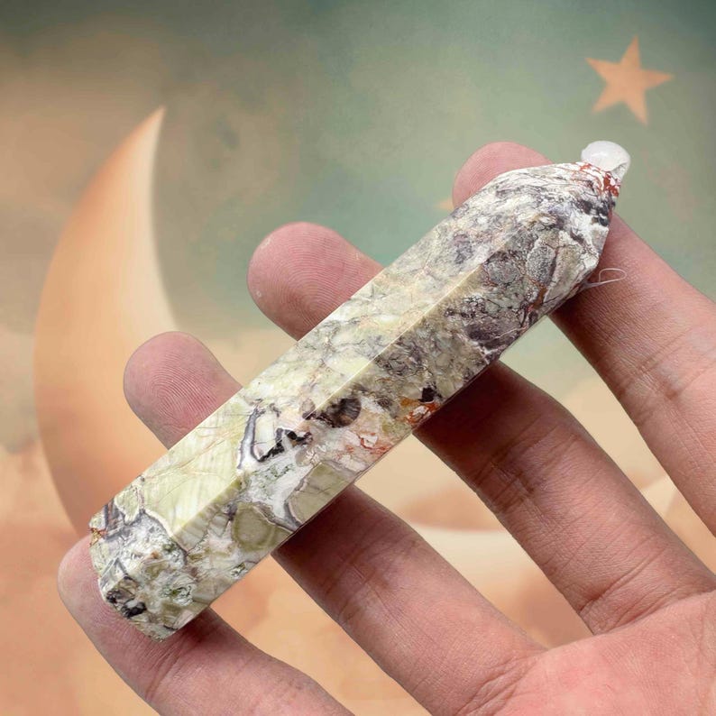 May include: A faceted, elongated crystal point with a marbled pattern of green, brown, and white. The crystal is held in a hand, showcasing its polished surface and natural variations. A small, clear crystal is at the top.