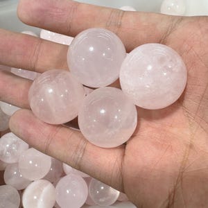 May include: A hand holds several polished, spherical rose quartz stones. The spheres are a soft pink color and have a smooth, translucent appearance. The background shows more of the same stones.