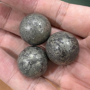 May include: Three spherical pyrite stones with a metallic, textured surface. The stones are a dark gray color with flecks of silver and gold, held in a hand. The overall appearance is of natural, raw gemstones.