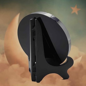 May include: A black plate stand, designed to hold a round object. The stand is made of plastic and has a hinged back for support. The background features a moon and star. The stand is suitable for plates or decorative items.