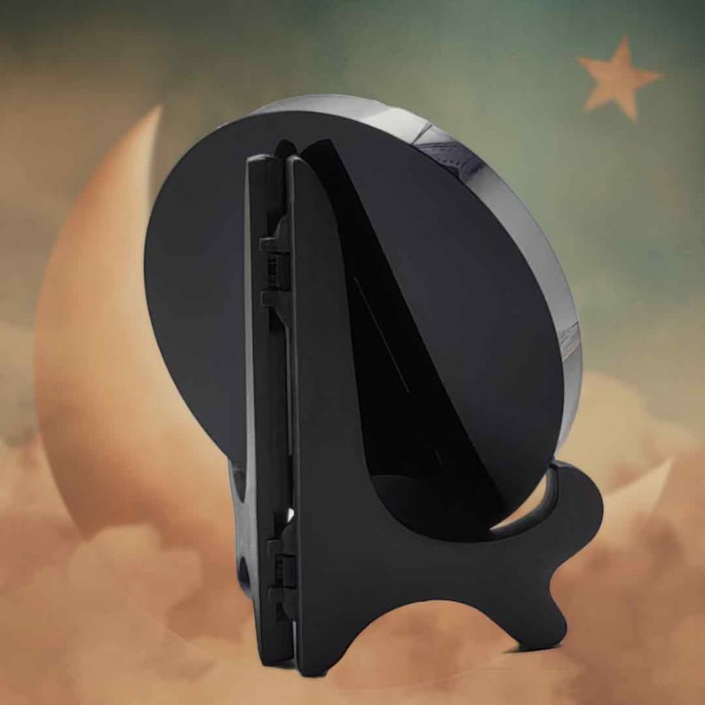 May include: A black plate stand, designed to hold a round object. The stand is made of plastic and has a hinged back for support. The background features a moon and star. The stand is suitable for plates or decorative items.