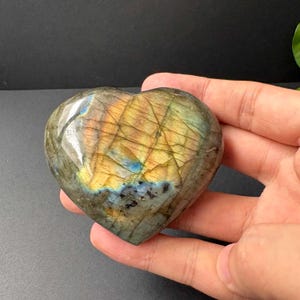 Puede incluir: Una piedra de labradorita en forma de corazón con una superficie pulida que presenta destellos iridiscentes en tonos dorados, azules y verdes. La piedra muestra fracturas internas naturales y una textura suave y redondeada.