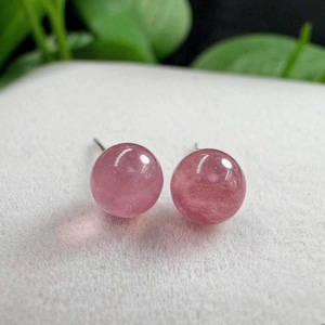 Puede incluir: Un par de pendientes de botón con cuentas esféricas y lisas de piedra preciosa rosa. Cada piedra redonda muestra una calidad translúcida con sutiles variaciones internas y una superficie pulida, unida a un poste de metal simple para sujeción.