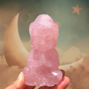 May include: A small, pink rose quartz Buddha statue. The Buddha is in a seated position with hands clasped in prayer. The statue is carved from a translucent pink stone. The background features a crescent moon and stars.