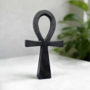 Könnte beinhalten: Ein dunkelgraues, metallisches Ankh-Symbol mit einer glatten, matten Oberfläche. Das Objekt hat eine kreisförmige Schlaufe an der Oberseite, einen horizontalen Querbalken und eine vertikale Basis, die sich nach unten hin leicht verjüngt.