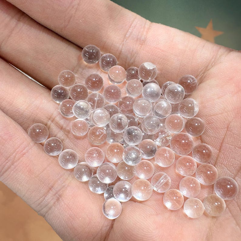 May include: A collection of small, clear, spherical beads with a slight pinkish-brown hue. The beads are clustered together, appearing translucent and smooth. The image showcases the beads in a close-up view, highlighting their round shape and subtle color variations.