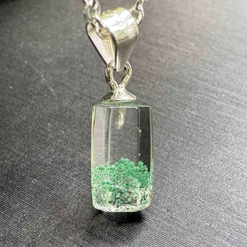 May include: A clear, rectangular pendant with a silver chain. The pendant contains a cluster of small, green, and white stones. The pendant is suspended from a silver bail.