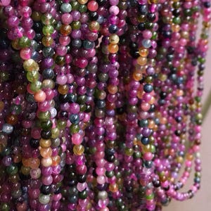 May include: Strands of colorful gemstone beads in shades of pink, purple, green, and brown. The beads are round and appear to be strung together, creating a textured and vibrant display. The image showcases a variety of colors and textures, perfect for jewelry making or crafting.