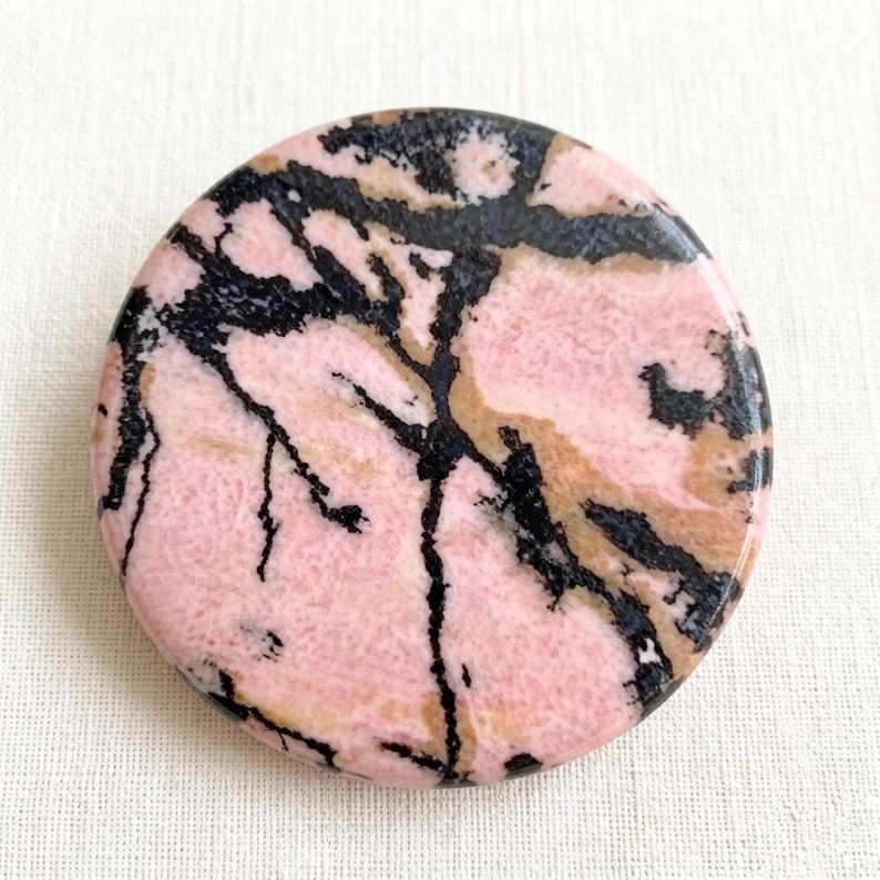 May include: A round, polished stone with a pink base color, featuring black and tan veining patterns. The stone's surface is smooth and reflective, showcasing a natural, organic design. The overall aesthetic is earthy and elegant.