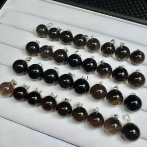 May include: A selection of spherical, dark brown gemstone pendants with silver-tone fixings. The pendants are displayed on a white surface, highlighting their polished, round form and diverse brown hues.