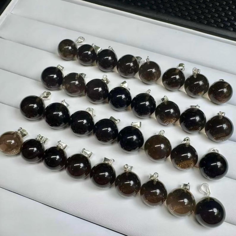 May include: A selection of spherical, dark brown gemstone pendants with silver-tone fixings. The pendants are displayed on a white surface, highlighting their polished, round form and diverse brown hues.
