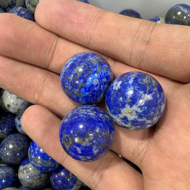 May include: Three polished lapis lazuli spheres with a deep blue hue and white and gold veining are held in a hand. The spheres are approximately 3.8 cm in diameter. Other spheres are visible in the background.
