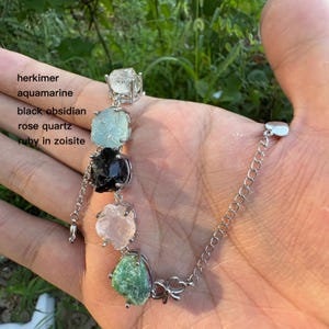 May include: A silver bracelet featuring a series of raw gemstones: herkimer, aquamarine, black obsidian, rose quartz, and ruby in zoisite. The bracelet is held in a hand, showcasing the natural colors and textures of the stones.