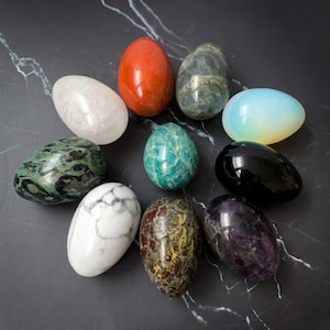 Puede incluir: Una colección de diez piedras pulidas en forma de huevo de varios colores y texturas, incluyendo rojo, blanco, negro, verde, morado y azul iridiscente, dispuestas en un patrón circular sobre una superficie oscura con vetas blancas.