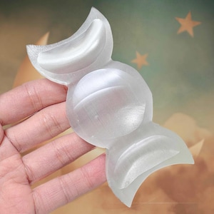 May include: A clear, selenite crystal carving in a stylized shape. The carving has a rounded central section with curved, wing-like extensions on either end. The crystal is held in a hand, with a soft, diffused background.