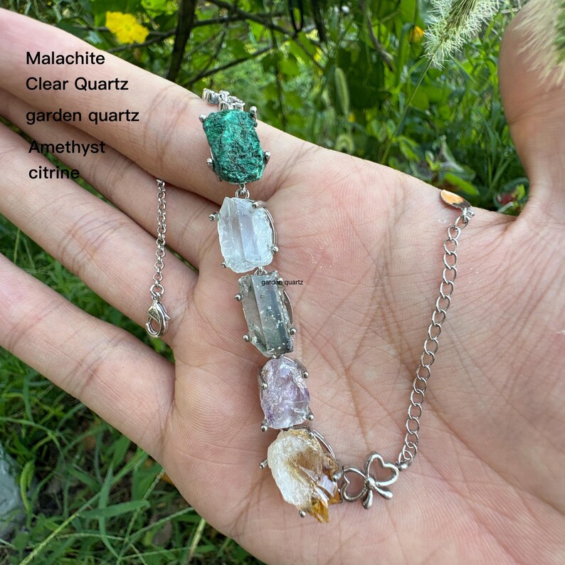 May include: A silver bracelet featuring a series of raw gemstones, including malachite, clear quartz, garden quartz, amethyst, and citrine. The gemstones are connected by silver links and a delicate chain with a lobster clasp. The bracelet is displayed against a natural background.