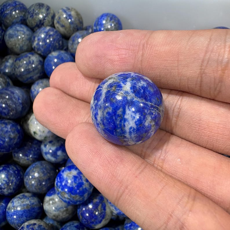 May include: A selection of spherical lapis lazuli stones. The main stone, held in a hand, is a deep blue with white and grey veining. Other stones are visible in the background.