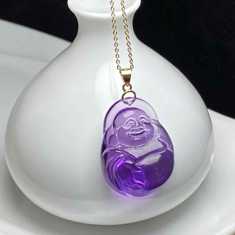 May include: A purple pendant necklace featuring a smiling Buddha figure. The pendant is carved from a translucent, amethyst-colored material and hangs from a delicate gold chain. The necklace is displayed against a white background.