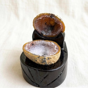 May include: Two halves of a geode, revealing a crystal interior with white and purple hues. The outer shell is tan and brown. The geodes are presented on a black, carved wooden stand, offering a striking contrast in colour and texture.
