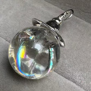 May include: A clear, spherical crystal pendant with a silver-coloured metal cap and loop. The crystal refracts light, displaying a rainbow of colours. The pendant is designed to be worn as a necklace or used as a decorative item.