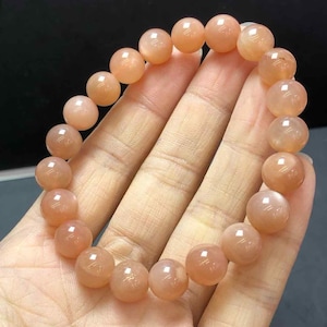 Op de afbeelding: Een perzik kleurige kralenarmband. De armband is gemaakt van ronde, gepolijste kralen met een subtiele glans. De kralen zijn aan elkaar geregen om een complete cirkel te vormen. De armband wordt in een hand gehouden.