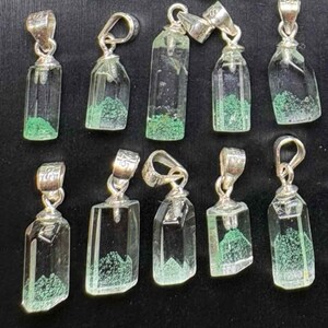May include: A collection of clear crystal pendants with silver-colored bails. Each pendant has a rectangular or irregular shape with a green, moss-like inclusion at the base. The pendants are displayed against a black background.