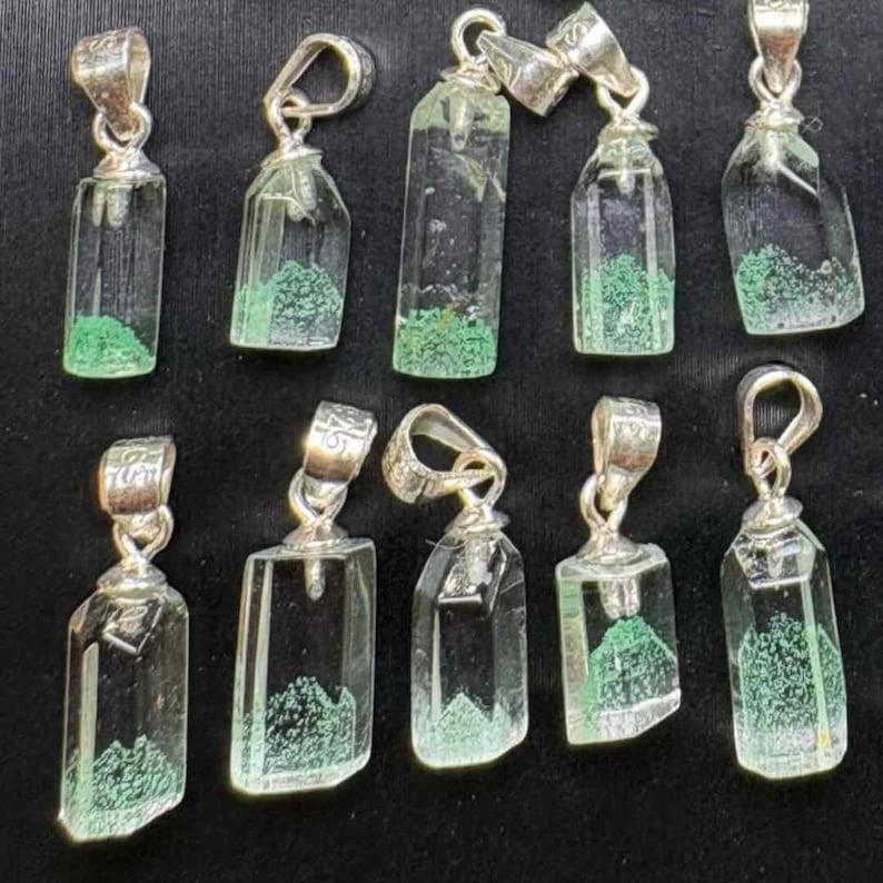 May include: A collection of clear crystal pendants with silver-colored bails. Each pendant has a rectangular or irregular shape with a green, moss-like inclusion at the base. The pendants are displayed against a black background.