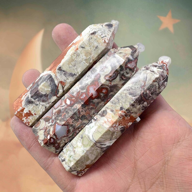May include: Four polished gemstone wands with a mix of colors including red, brown, cream, and gray. Each wand has a pointed tip and a faceted, rectangular shape. The wands are held in a hand.