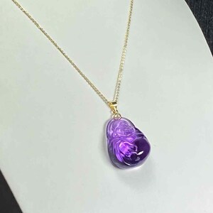 May include: A gold-colored chain necklace with a pendant. The pendant is a carved, purple gemstone with a detailed design. The gemstone is a deep, rich purple color, and the chain is delicate and shiny.