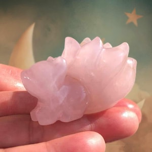 May include: A carved pink quartz figurine of a lotus flower. The sculpture is held in a hand, showcasing the delicate details of the petals. The background features a soft, celestial design with a crescent moon and a star.