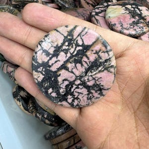May include: A round, polished stone with a pink and black pattern. The stone is held in a hand, with other similar stones visible in the background. The pink color is dominant, with black veining throughout. The stone appears to be a decorative item.