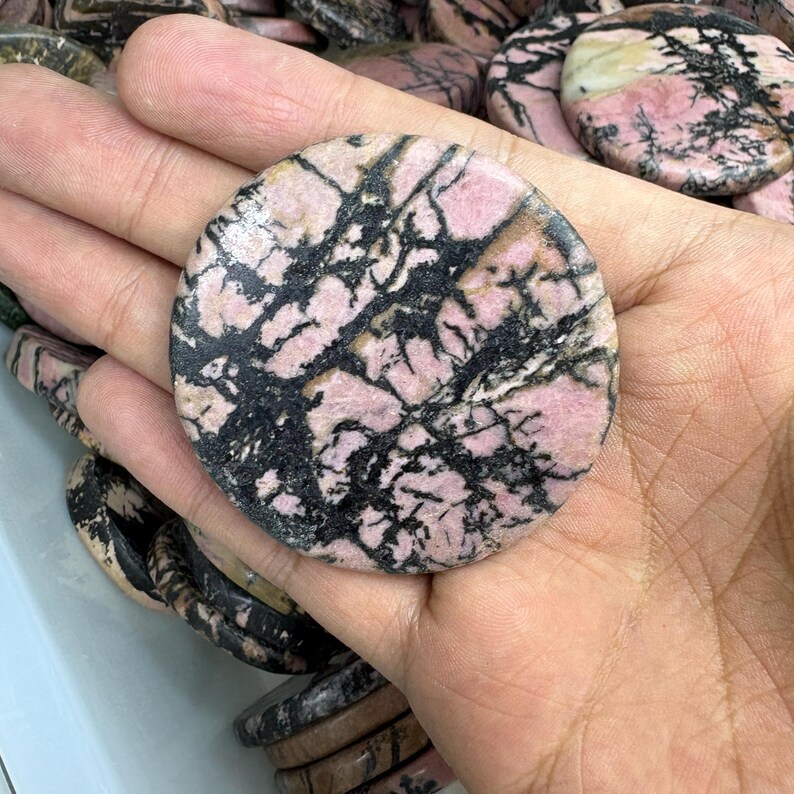 May include: A round, polished stone with a pink and black pattern. The stone is held in a hand, with other similar stones visible in the background. The pink color is dominant, with black veining throughout. The stone appears to be a decorative item.