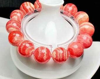 May include: A beaded bracelet featuring large, round beads in shades of red and cream. The beads have a marbled appearance and are strung together. The bracelet is displayed around a white, ceramic vase. The background is dark.