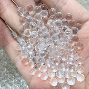 May include: A handful of clear, spherical crystal beads. The beads are translucent with a slight pinkish hue, and vary in size. The image is a close-up, showing the beads filling the palm of a hand.