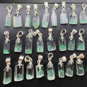 May include: Collection of clear glass pendant necklaces with silver-colored metal accents. Each pendant has a unique shape and contains a green, textured material, resembling a miniature landscape. The pendants are displayed against a dark background.