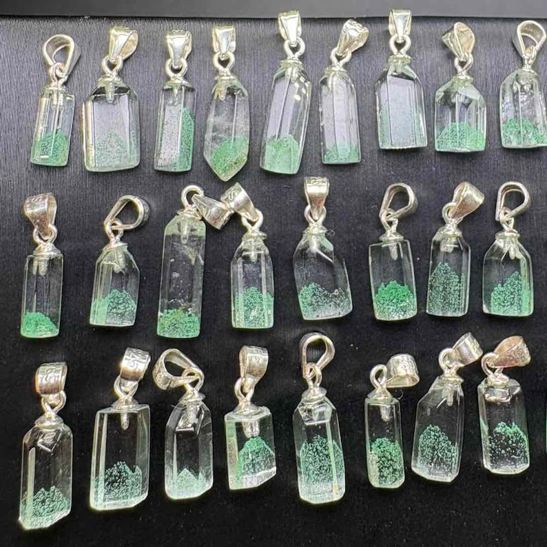 May include: Collection of clear glass pendant necklaces with silver-colored metal accents. Each pendant has a unique shape and contains a green, textured material, resembling a miniature landscape. The pendants are displayed against a dark background.