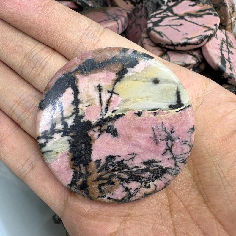 May include: A round, polished gemstone with a pink base color, accented by black and white veining. The stone has a natural, organic pattern resembling a landscape. The gemstone is held in a hand, showcasing its size and texture. This item is ideal for jewelry making or collecting.