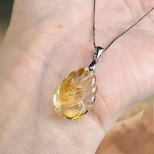 May include: A pendant necklace with a carved, teardrop-shaped gemstone in shades of yellow and clear. The pendant hangs from a delicate silver chain. The gemstone has intricate details and is held in a silver setting.