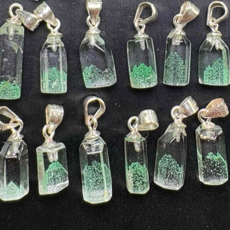 May include: A collection of clear glass pendant necklaces with silver-colored metal findings. Each pendant has a bottle shape with a green, textured interior. The pendants are displayed against a dark background.