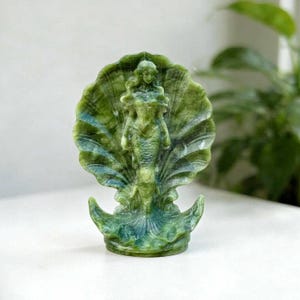 May include: A carved green stone figurine depicting a mermaid standing in front of a large, open scallop shell. The sculpture features variegated shades of olive, forest green, and teal, with a smooth, polished finish and intricate detailing.
