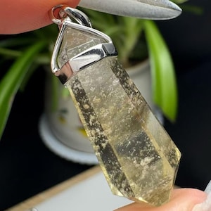May include: A faceted, light yellow crystal pendant with a silver-colored bail. The crystal has a slightly cloudy appearance and is held by a silver-colored metal setting. The pendant is held by a person's fingers.