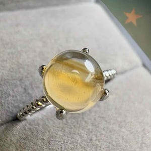 Könnte beinhalten: Ein Silberring mit einem großen, klaren, runden Edelstein mit goldgelben Einschlüssen. Der Edelstein wird von vier silbernen Zinken gehalten. Das Band des Rings hat ein gedrehtes Design.