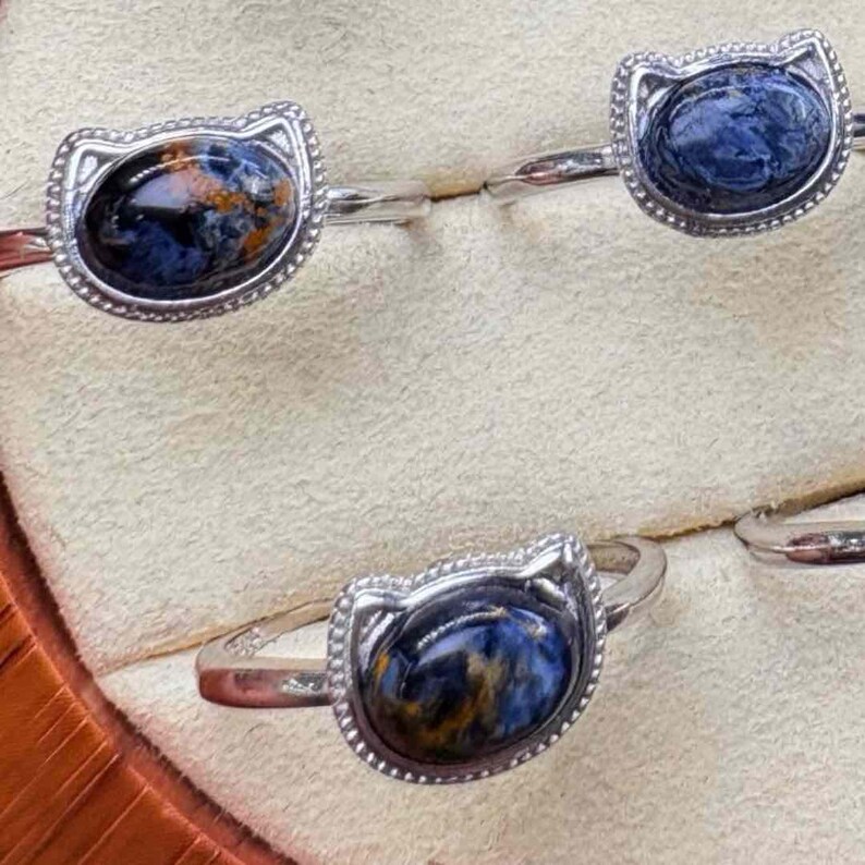 May include: Silver rings with cat-shaped settings, each featuring a polished, oval gemstone. The gemstones display a mix of blue, brown, and orange hues. The rings are displayed on a light-colored surface.