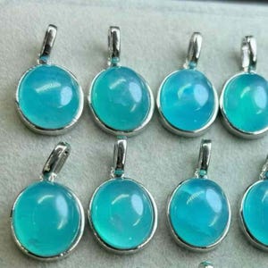 May include: A selection of oval pendants featuring a vibrant, translucent teal gemstone set in silver-coloured metal. Each pendant has a small loop at the top for a chain. The pendants are closely arranged, indicating a jewellery collection.